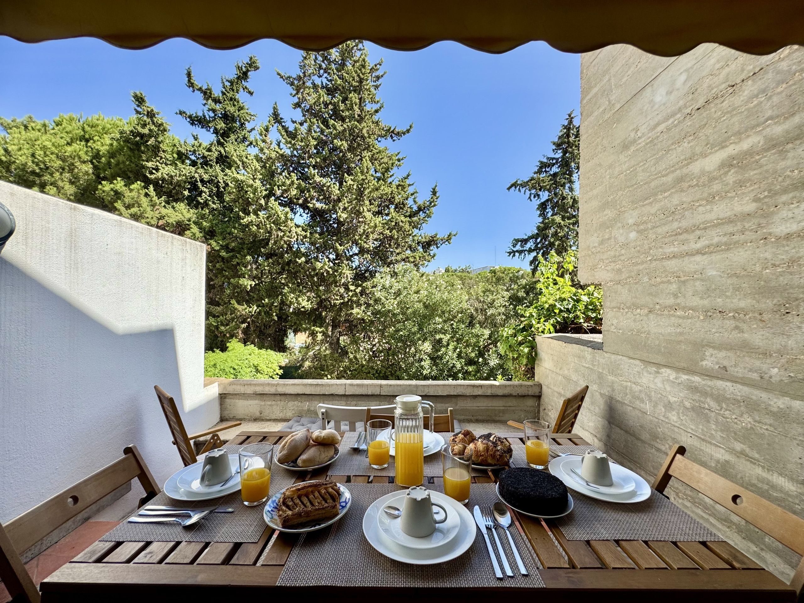 An outdoor dining area with a table set for breakfast, including bread, pastries, and a cake, with a view of trees and a clear sky.