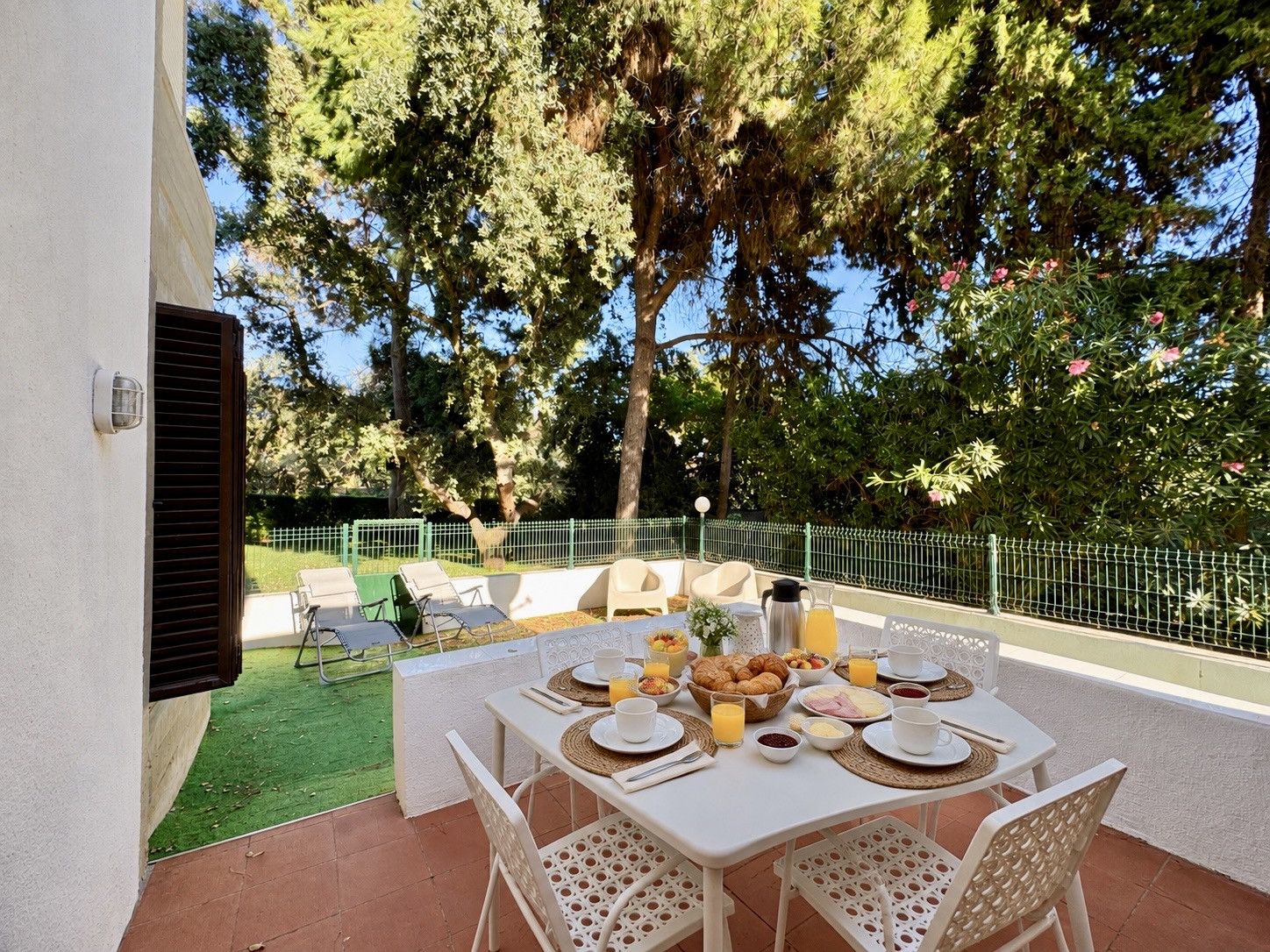 An outdoor dining area with a table set for breakfast, surrounded by chairs, and a view of a garden with trees and a fence.