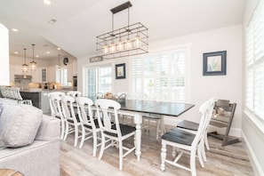 A modern kitchen with white cabinets, a dining area with a black table and white chairs, and a large window with blinds. - Spacious 8BR Dewey Beach Home | Private Seasonal Pool + Decks (Dewey Beach)