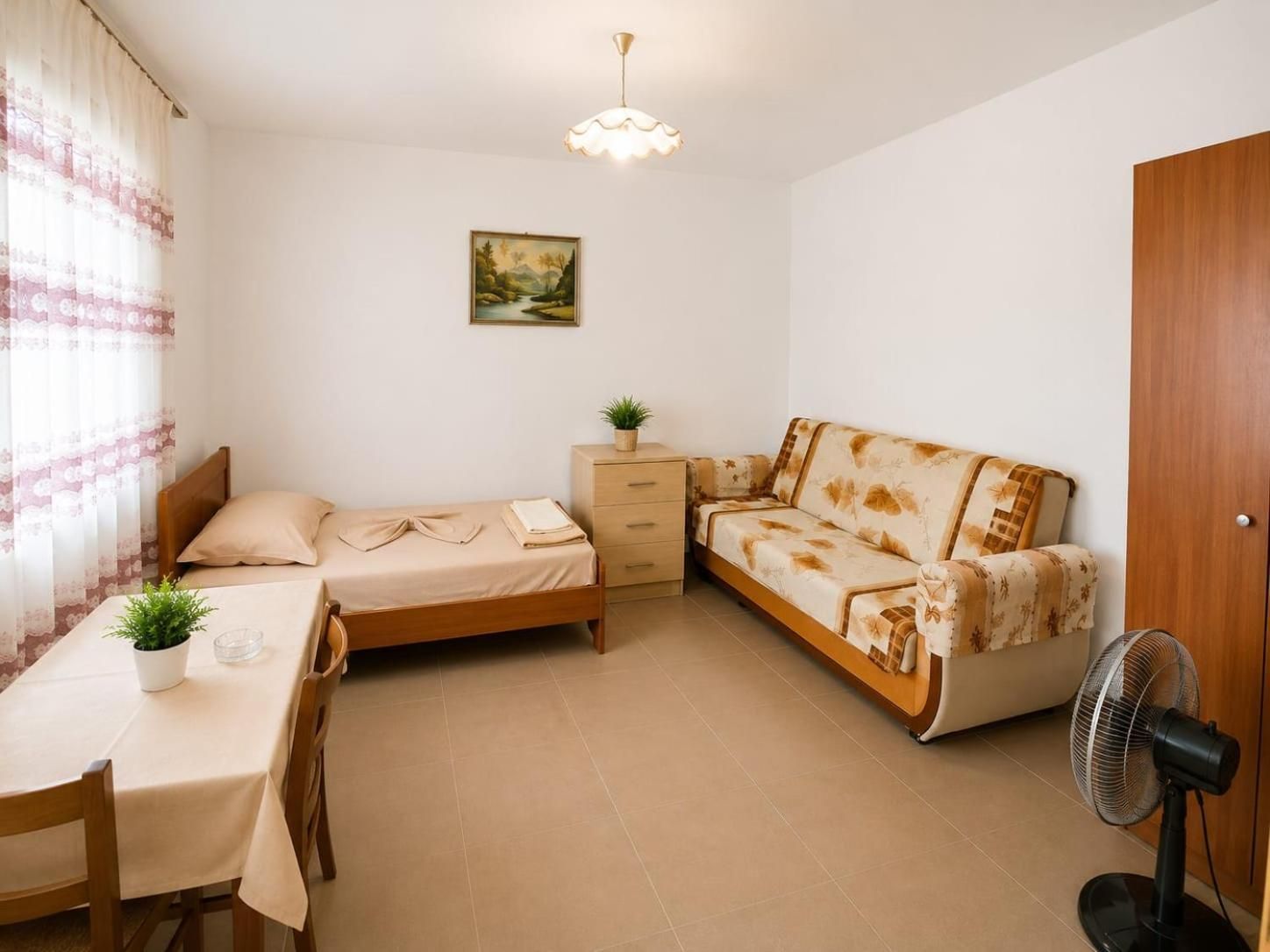 A small, well-lit room with a bed, a desk, a chair, a bedside table with a plant, a sofa, and a standing fan.