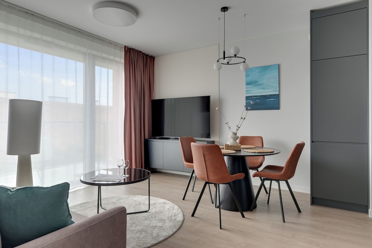 Ein modernes Wohnzimmer mit einer Couch, einem Esstisch mit Stühlen, einem Fernseher und einem großen Fenster mit Vorhängen.