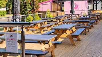 Un área de estar al aire libre con mesas de picnic de madera y sombrillas, un letrero de precaución sobre una cubierta resbaladiza, y un edificio rosa al fondo.