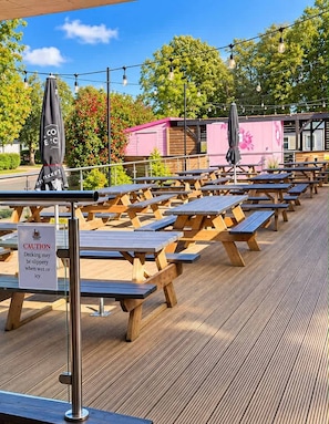 Ein Sitzbereich im Freien mit Holztischen, Sonnenschirmen, einem Warnschild wegen rutschiger Terrassenplatten und einem rosafarbenen Gebäude im Hintergrund.