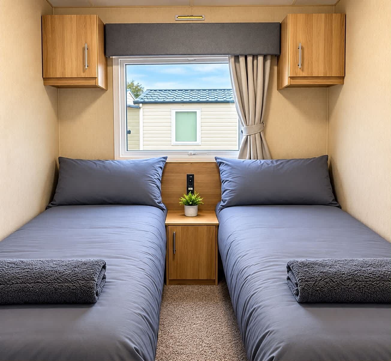 A compact RV bedroom with two beds, a small table with a plant, and a window with curtains.