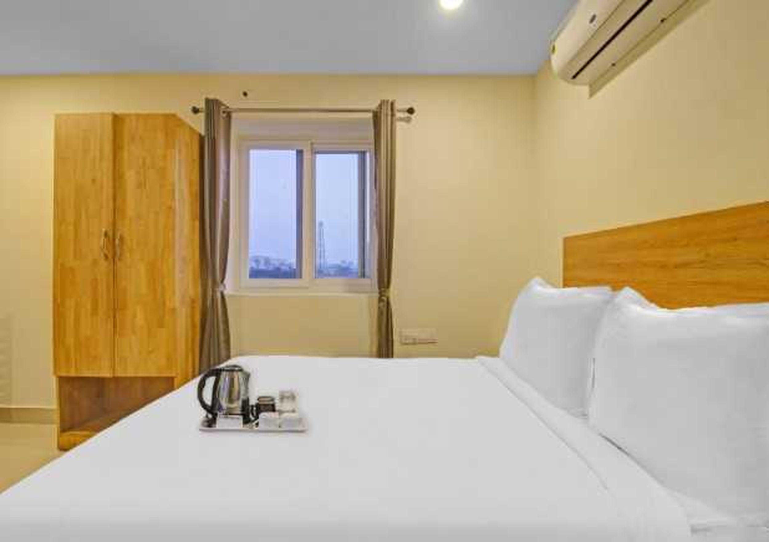 A hotel room with a bed, a wooden wardrobe, a window with curtains, and a bedside table with a kettle and a cup.