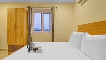 A hotel room with a bed, a wooden wardrobe, a window with curtains, and a bedside table with a kettle and a cup.