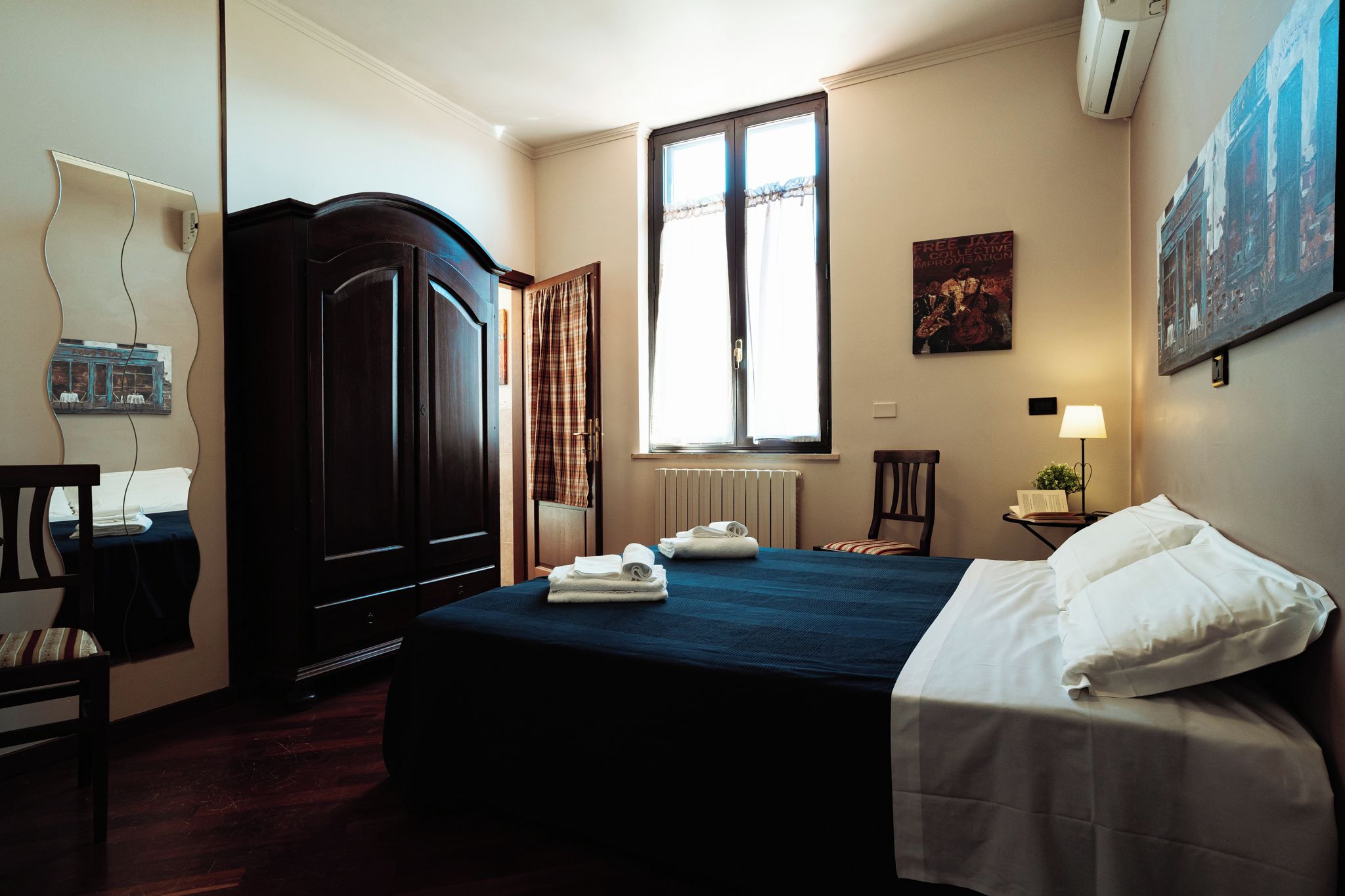 Una habitación de hotel con cama, armario, ventana con cortinas, radiador, sillón, escritorio con lámpara y aire acondicionado instalado en la pared.