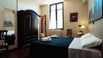 Una habitación de hotel con cama, armario, ventana con cortinas, radiador, sillón, escritorio con lámpara y aire acondicionado instalado en la pared.