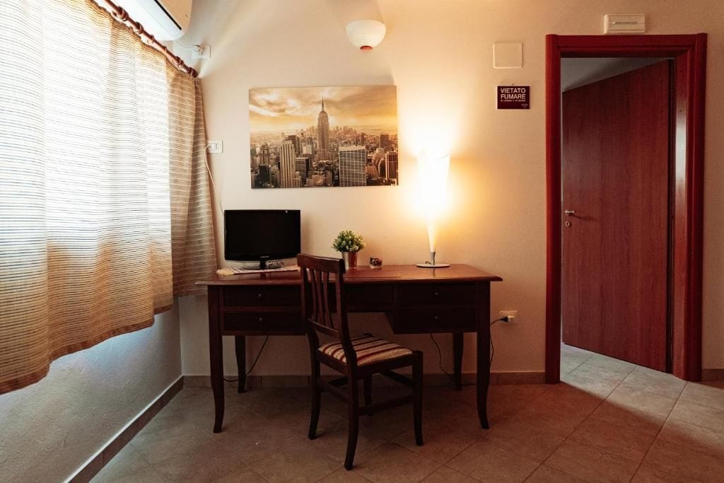 Una habitación con escritorio, silla, televisor de pantalla plana, lámpara de pared y puerta con un cartel.