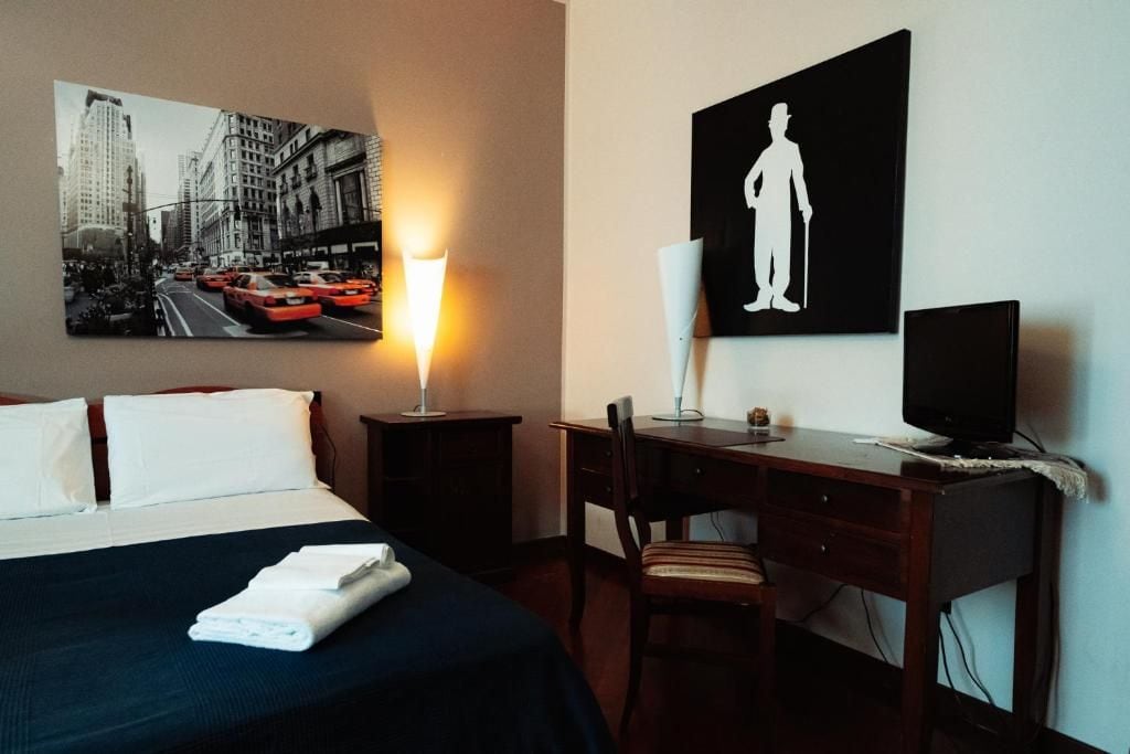 A hotel room with a bed, a desk, a chair, a television, a lamp, and a painting of a cityscape.