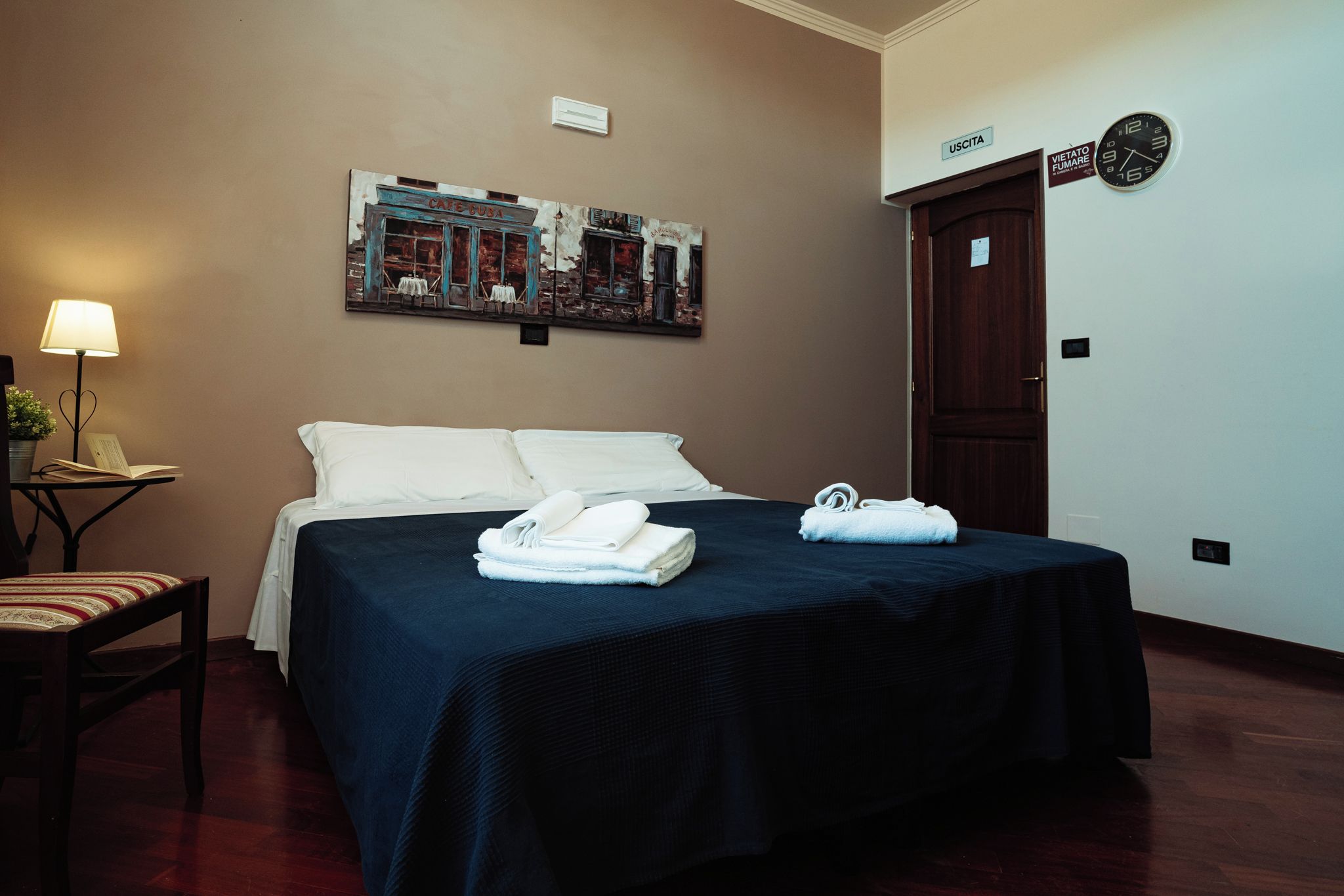 Habitación de hotel con una cama, un escritorio con lámpara, un reloj de pared y una puerta con la etiqueta 'USCITA'.