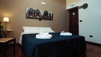 Habitación de hotel con una cama, un escritorio con lámpara, un reloj de pared y una puerta con la etiqueta 'USCITA'.