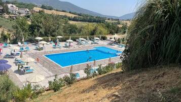 Zona de piscina con sillas de descanso y sombrillas, rodeada de vegetación y montañas al fondo.