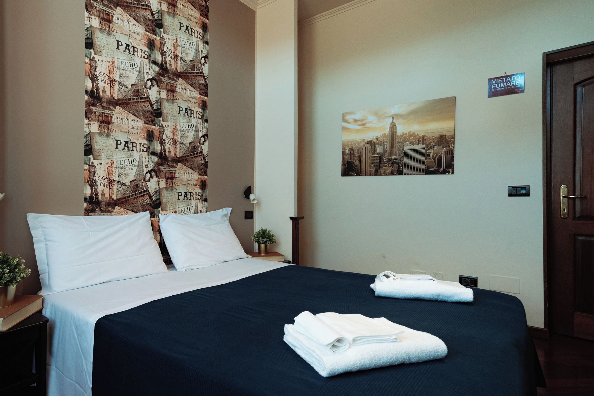 Una habitació d'hotel amb un llit, almoines, una taula de nit, una paret amb un quadre de paisatge urbà i una porta.