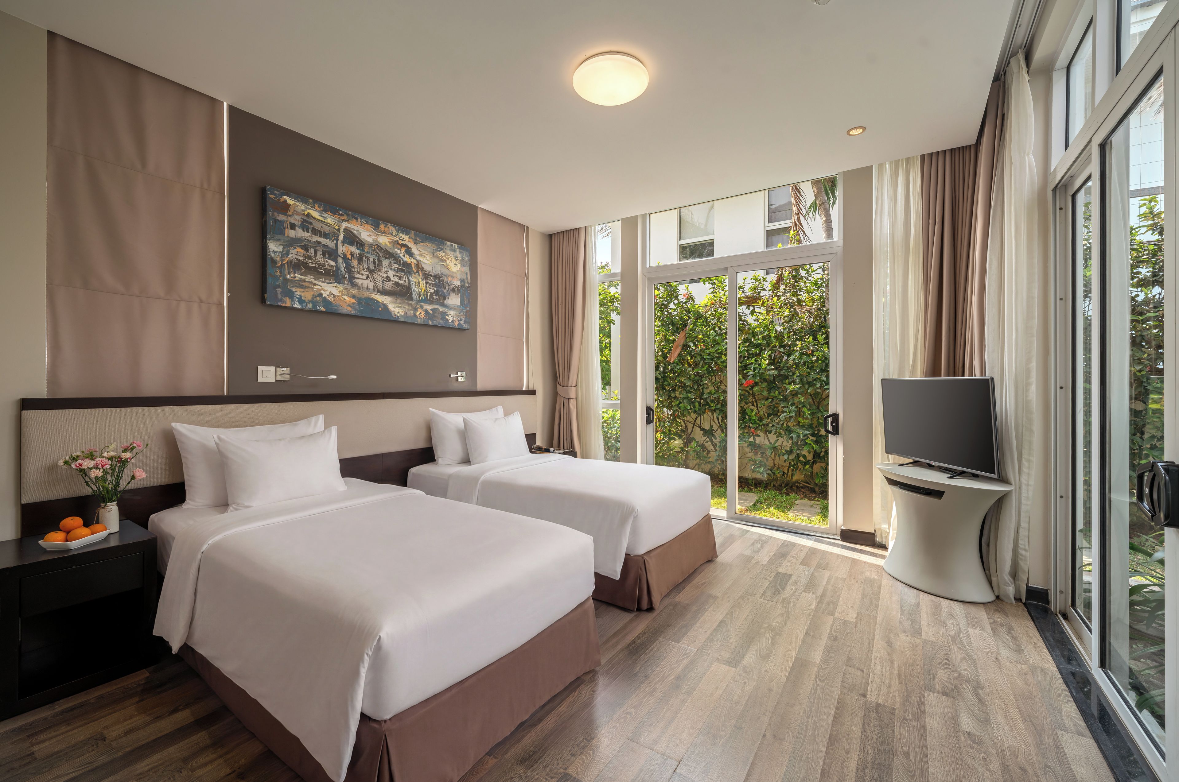 Quarto de hotel com duas camas, televisão e uma janela grande com vista para vegetação.