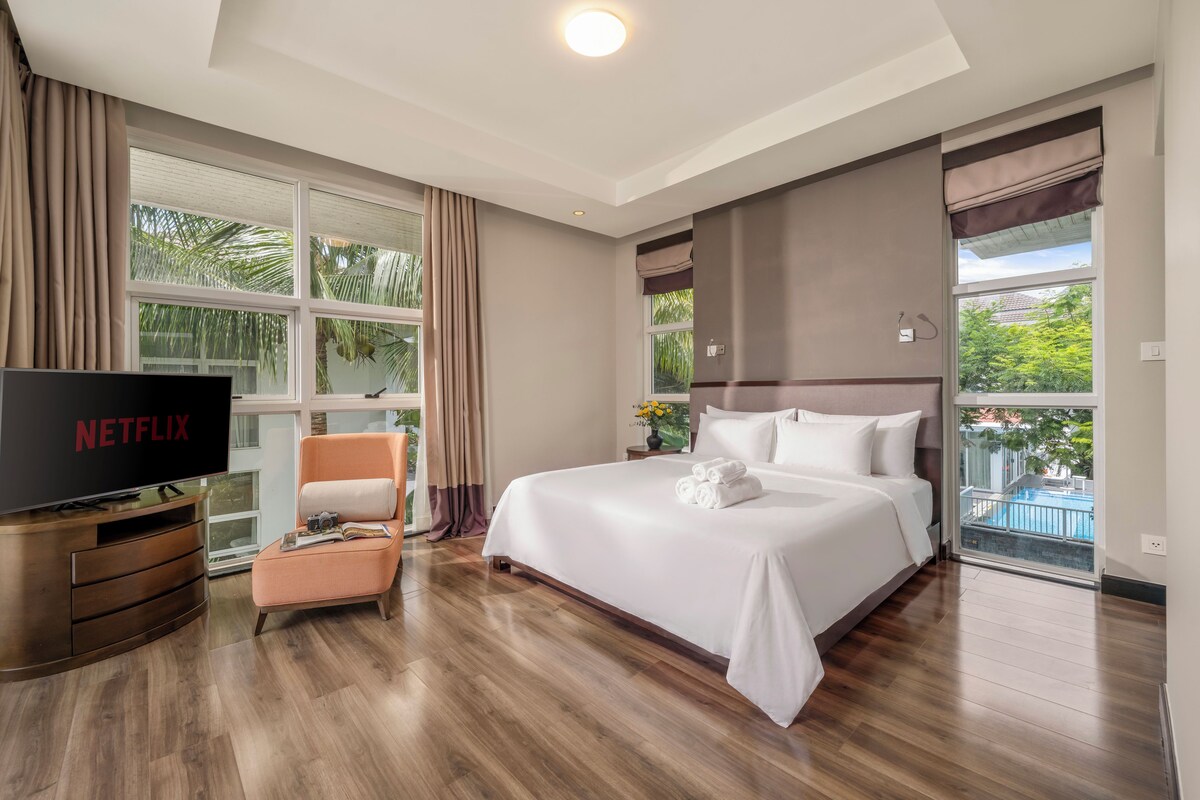 Ein Hotelzimmer mit einem großen Bett, einem Fernseher, einer Sitzecke und Blick auf einen Pool.