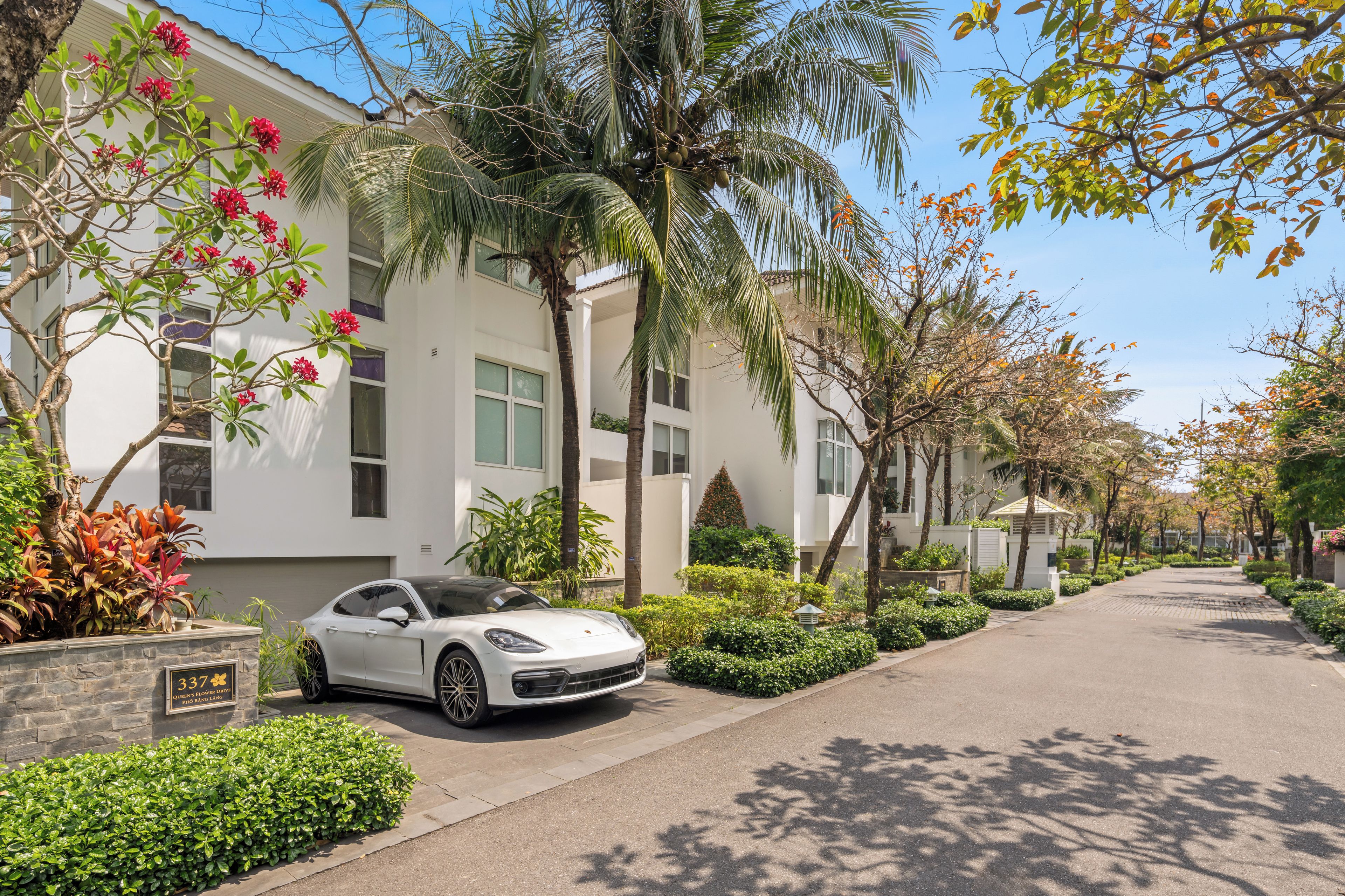 Um carro branco estacionado numa rua arborizada e limpa, com um edifício de grandes janelas e um jardim bem cuidado.