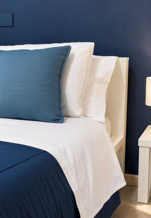 Una cama con ropa de cama blanca y azul, una mesita de noche con una lámpara y una pared azul oscuro.