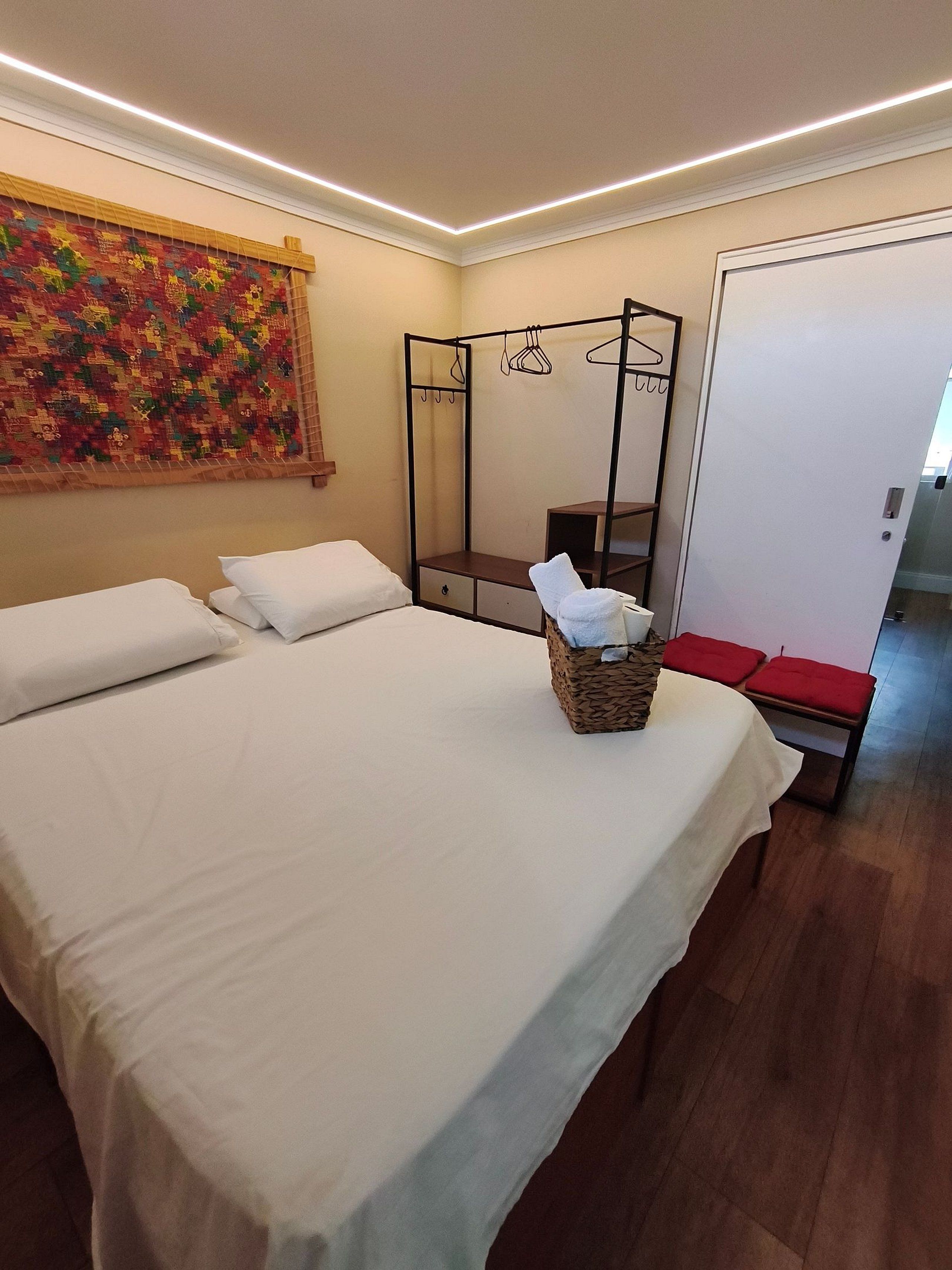 A hotel room with a large bed, a bedside table, a hanging coat rack, a wardrobe, and a colorful tapestry on the wall.
