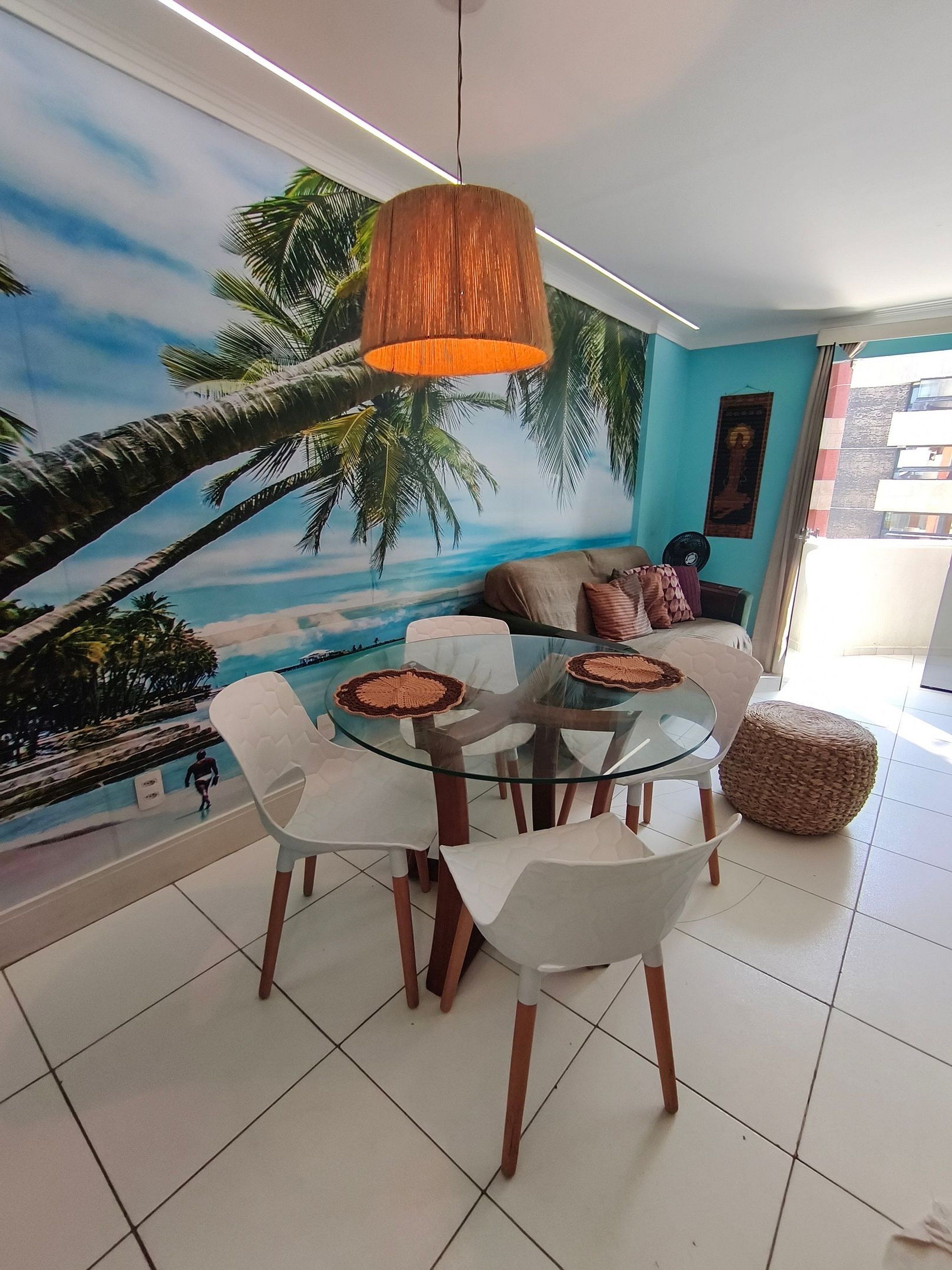 A dining area with a glass table and chairs, a hanging lamp, and a mural of a beach scene on the wall.