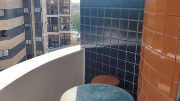 A small balcony with a green table and a brown stool, a tiled wall, and a view of a building with multiple windows.