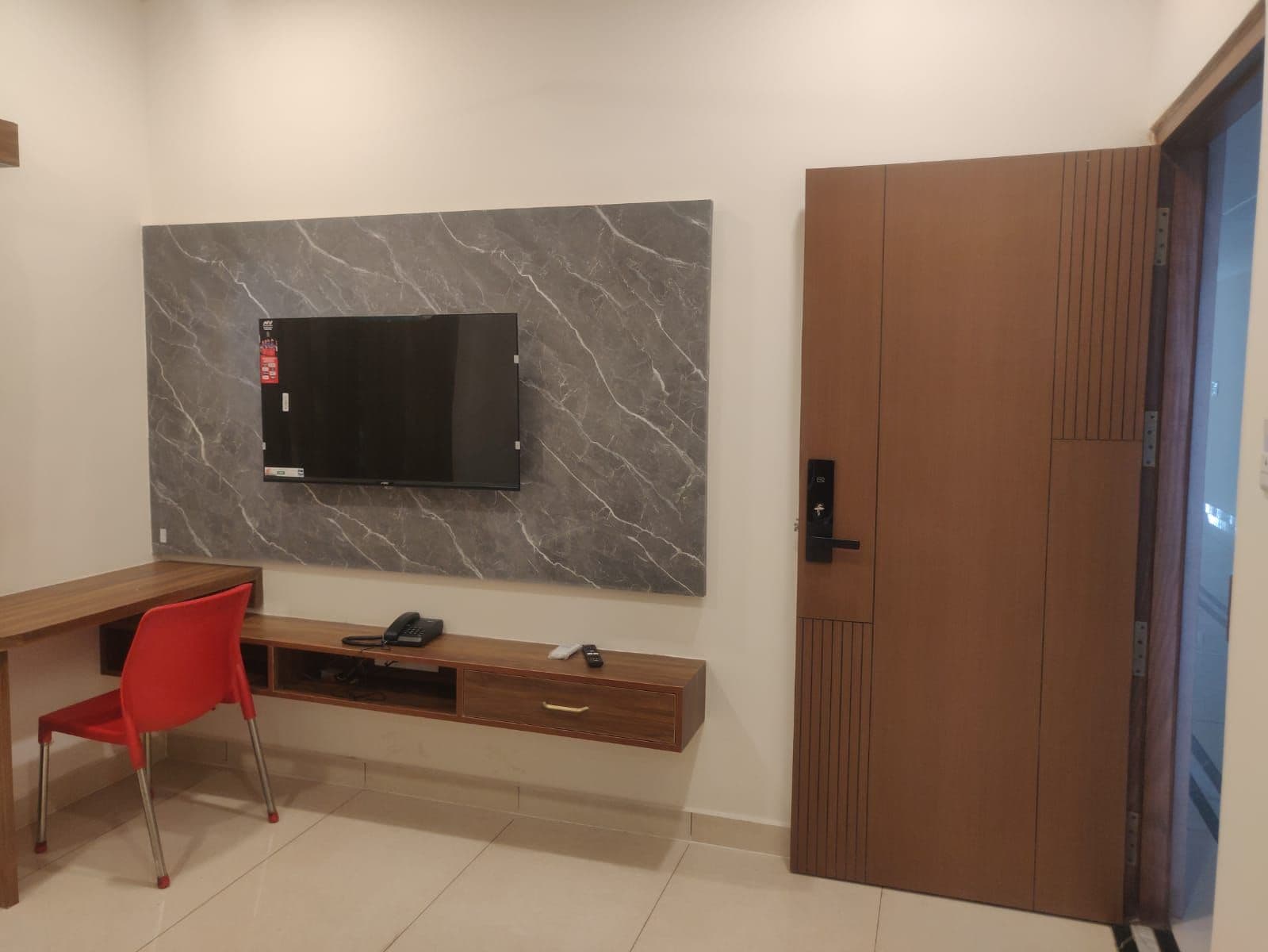 Une chambre moderne avec un téléviseur à écran plat fixé sur un mur de marbre, un bureau en bois avec une chaise rouge et une porte coulissante en bois.