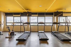 A gym with treadmills, stationary bikes, and a rowing machine, large windows with a city view, and a yellow ceiling. - ArchHome by Bhomy (São Paulo)