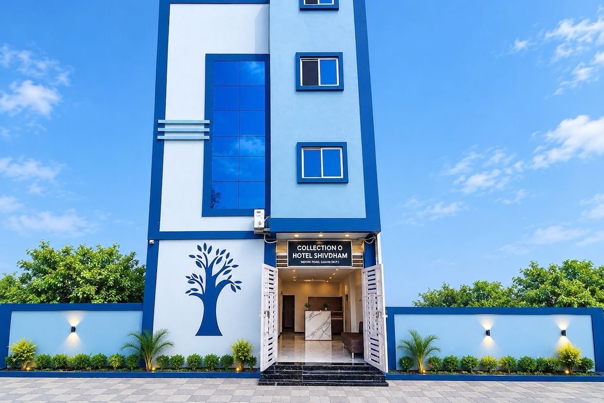 A modern hotel with a blue and white facade, a tree mural, and a sign for 'Collection O Hotel Shivdham.'