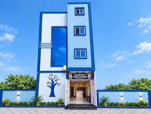 Ein modernes Hotel mit blauer und weißer Fassade, einer Baum-Mural und einem Schild für 'Collection O Hotel Shivdham'.