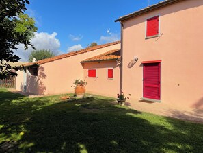 A building with a red door and window shutters, a large potted plant, and a clear blue sky. - Charming country home with a pool and a large fenced-in yard (Cortona)