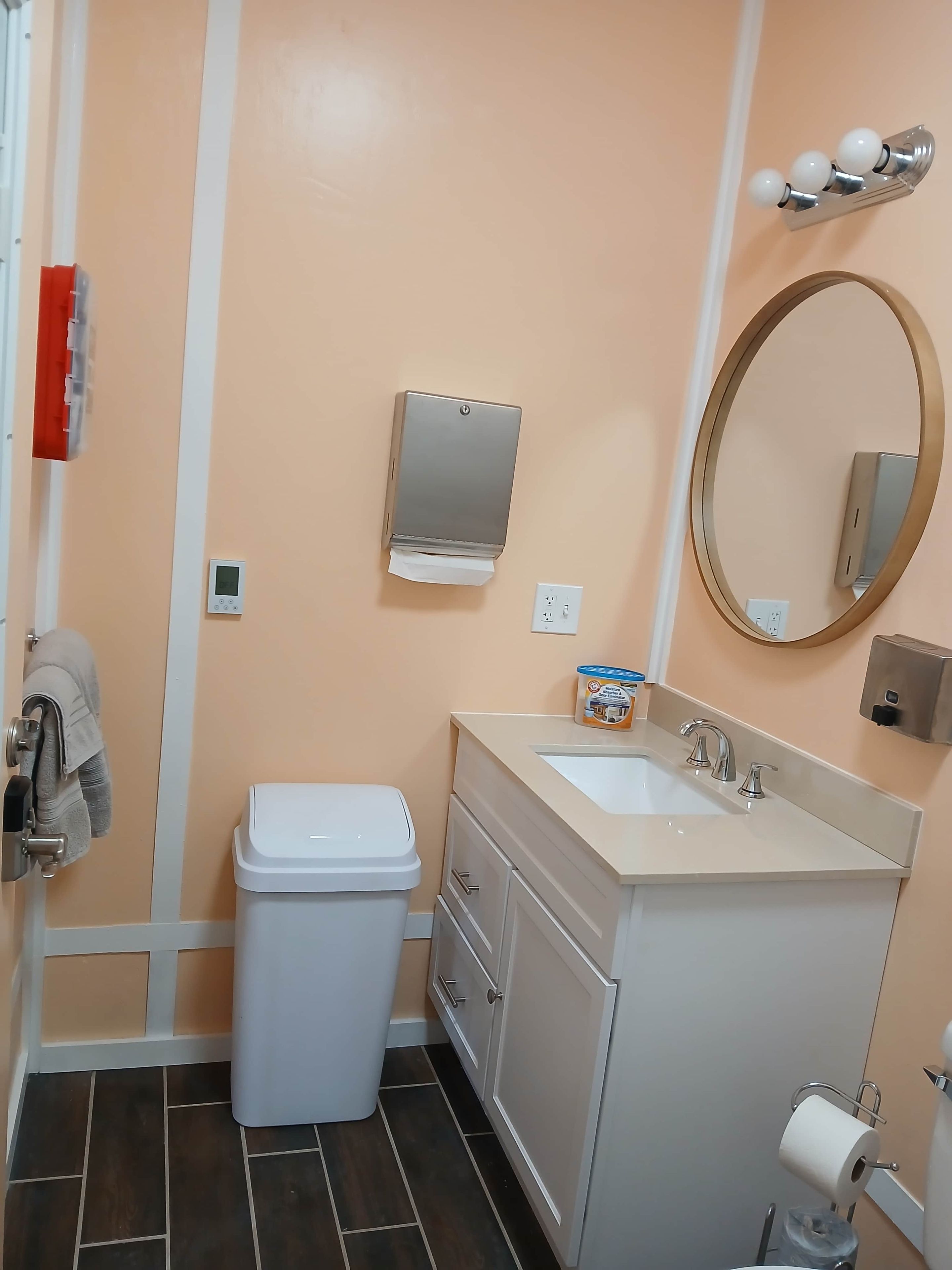 A bathroom with a toilet, sink, mirror, towel dispenser, and a trash bin.