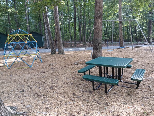 Une aire de jeux avec une structure d’escalade et des tables de pique-nique, située dans un espace boisé.