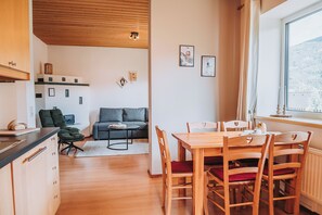 Un intérieur moderne avec une table à manger en bois, un canapé gris et un coin cuisine doté de placards et d’une fenêtre.