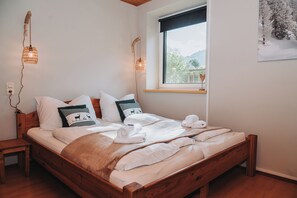 Un lit en bois avec une literie blanche et des coussins décoratifs, une table de chevet, un luminaire fixé au mur et une fenêtre donnant sur un paysage verdoyant.