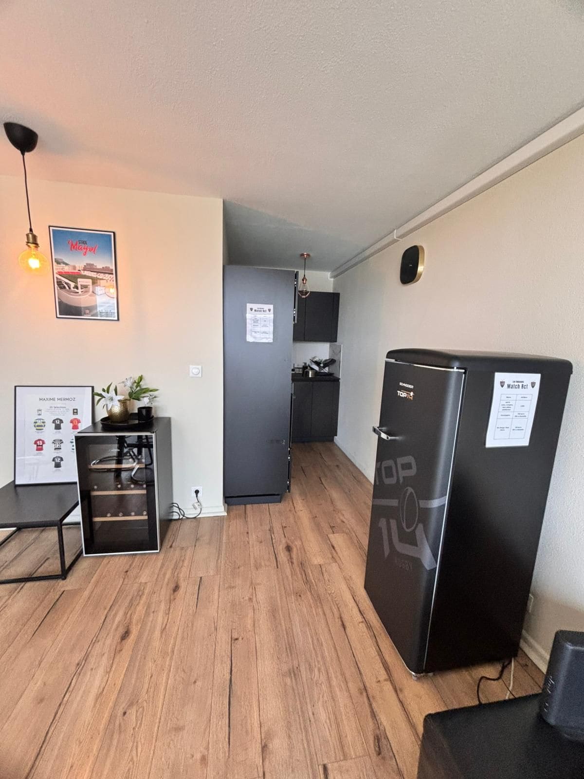 A compact living space with a kitchenette, a refrigerator, a dining table, and a wall-mounted light fixture.