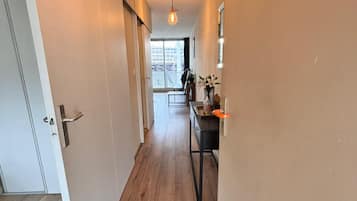 A hallway with wooden flooring, a door on the left, and a kitchen area with a counter and cabinets in the background.