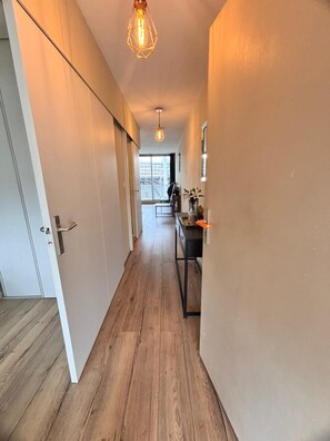 A hallway with wooden flooring, a door on the left, and a kitchen area with a counter and cabinets in the background. - Vrbo Property (Toulon)