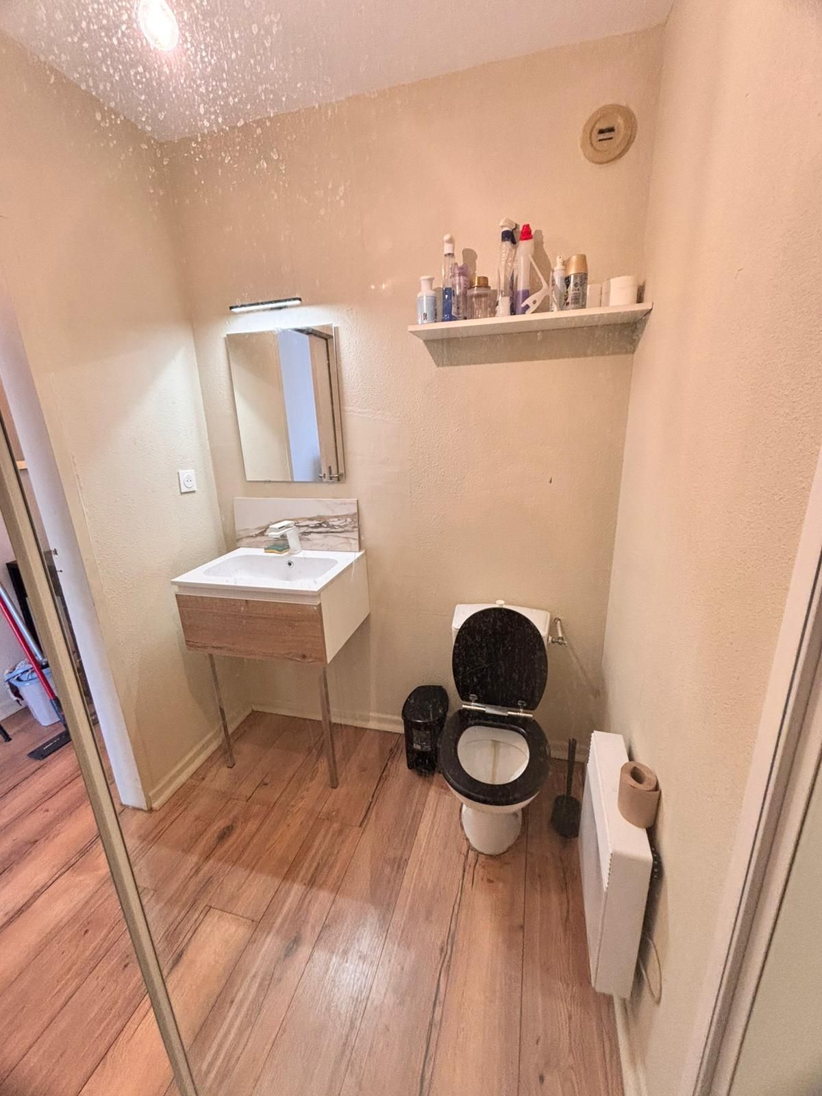 Une petite salle de bain avec un plancher en bois, un lavabo blanc, des toilettes noires et une étagère contenant des articles de toilette.