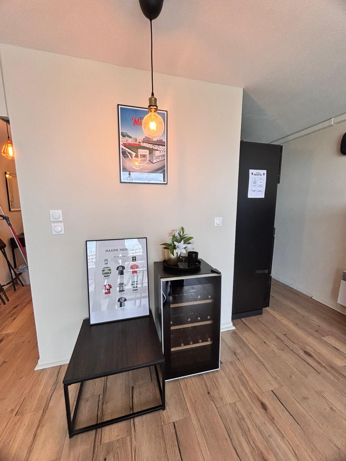 A room with a wooden floor, a black cabinet with drawers, a framed poster, a hanging light fixture, and a door with a sign.
