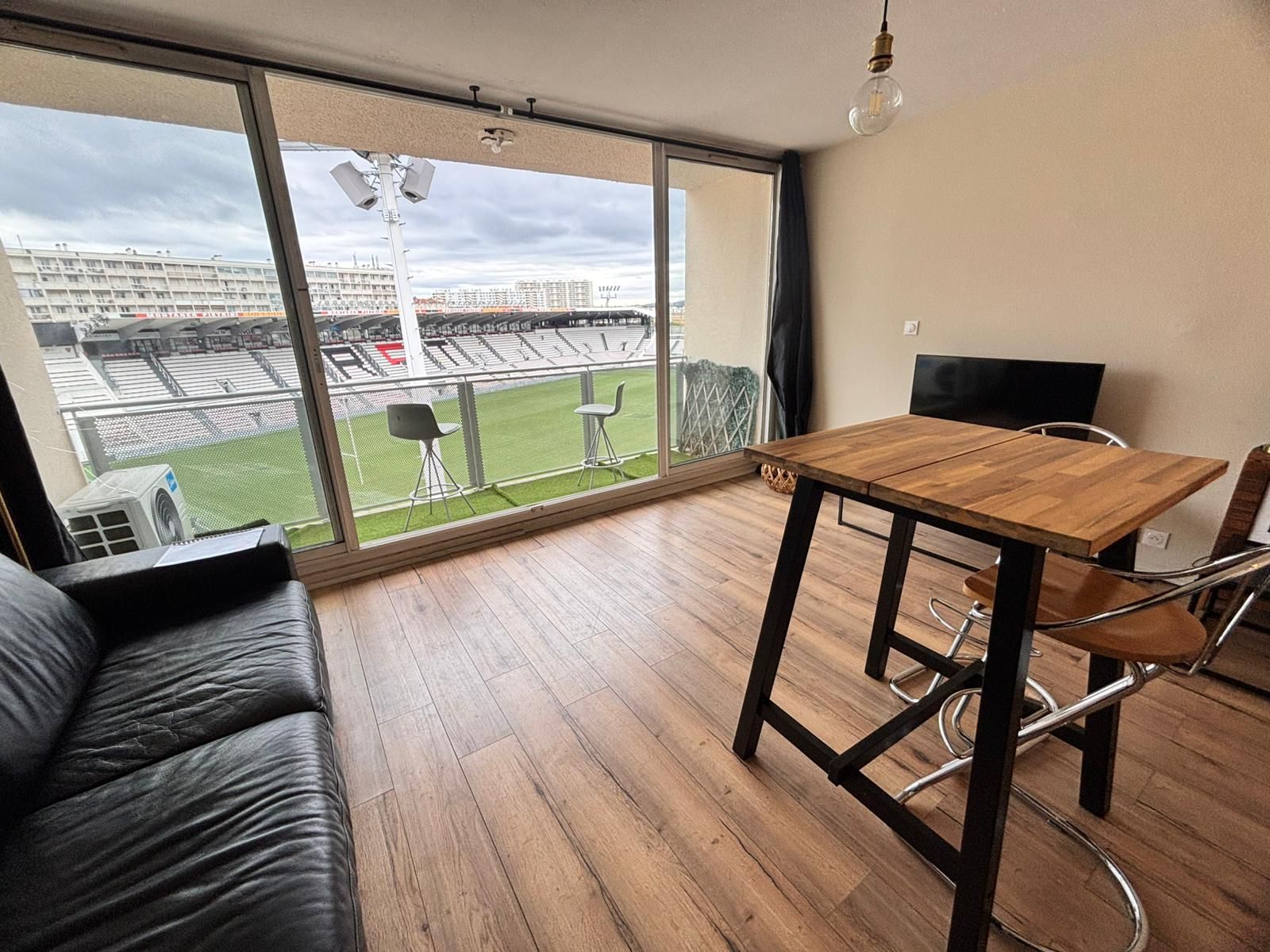 A room with a large window, a wooden table, a black leather sofa, and a television.