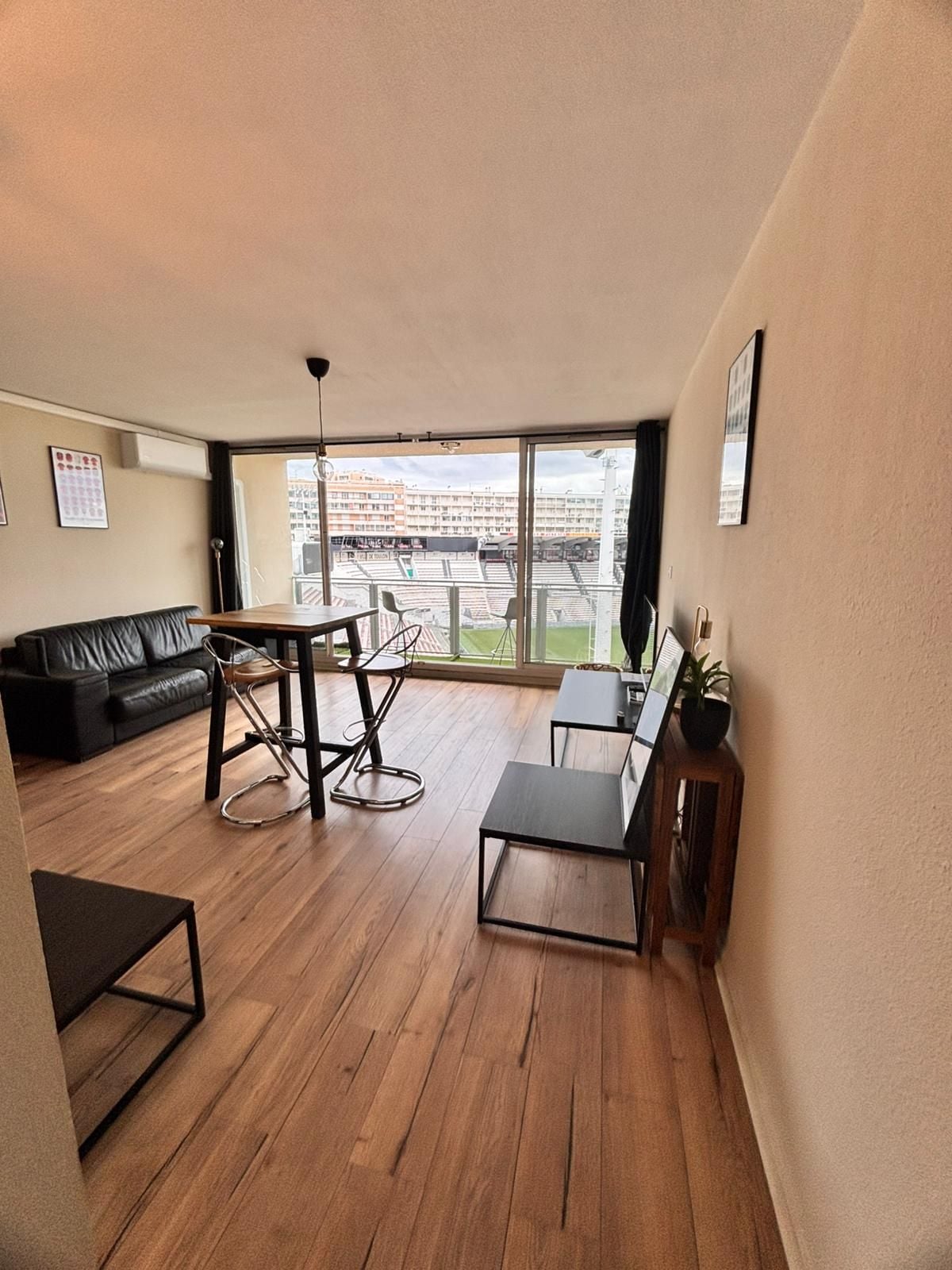A spacious living area with a wooden floor, a black sofa, a dining table with chairs, and a large window offering a view of a stadium.