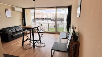 A spacious living area with a wooden floor, a black sofa, a dining table with chairs, and a large window offering a view of a stadium.