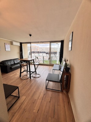 A spacious living area with a wooden floor, a black sofa, a dining table with chairs, and a large window offering a view of a stadium. - Vrbo Property (Toulon)