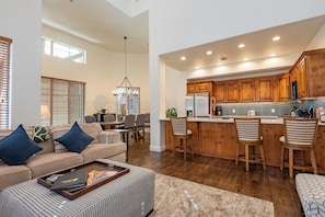 A spacious living area with a sofa, ottoman, and a coffee table with magazines. There is a dining area with a table and chairs, and a kitchen with wooden cabinets and a stainless steel refrigerator. - Central Park 22- New Listing! Luxury Condo Great Location Close to Ski Lift (Ketchum)