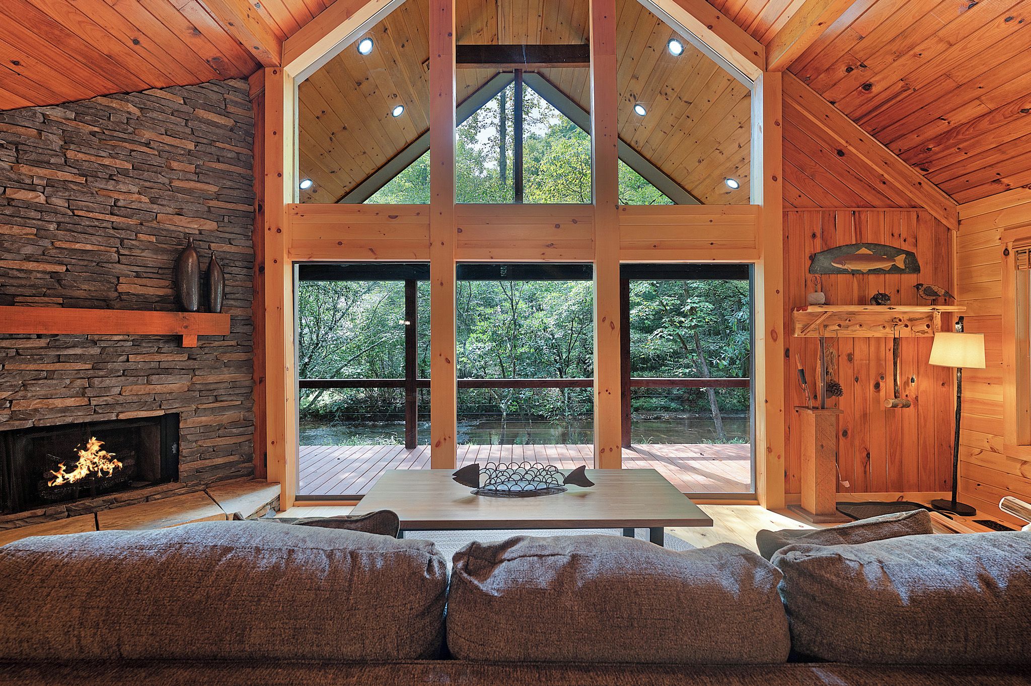 A log cabin interior with a stone fireplace, large windows, a wooden deck, and a cozy seating area.