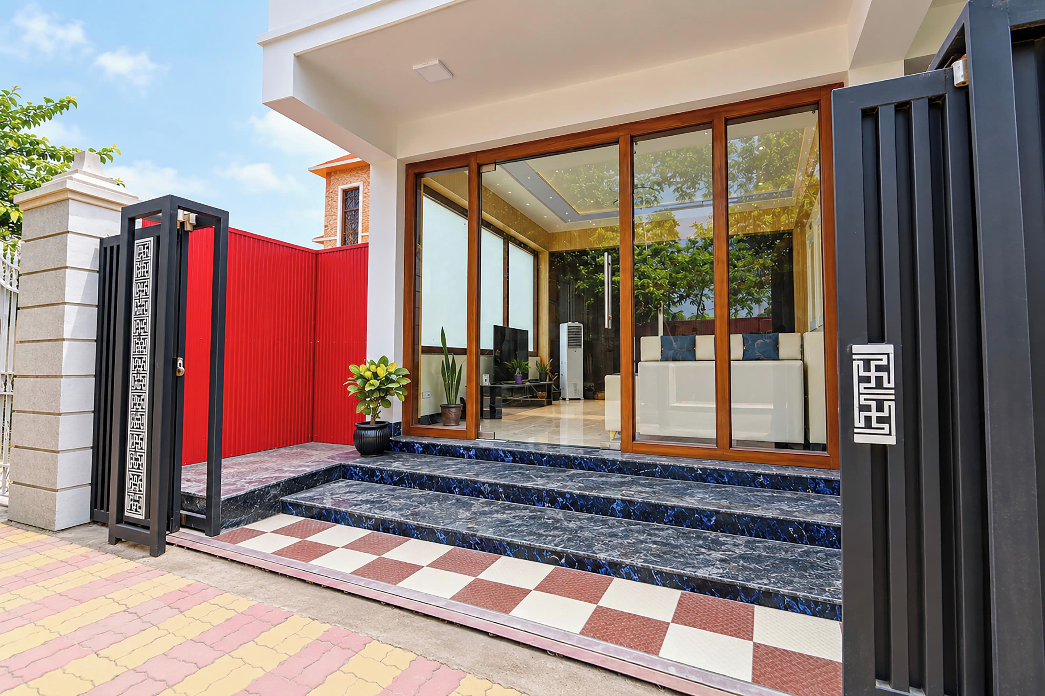 A modern house with a red gate, a tiled entrance, and a large glass door leading to a living area with a sofa and a plant.