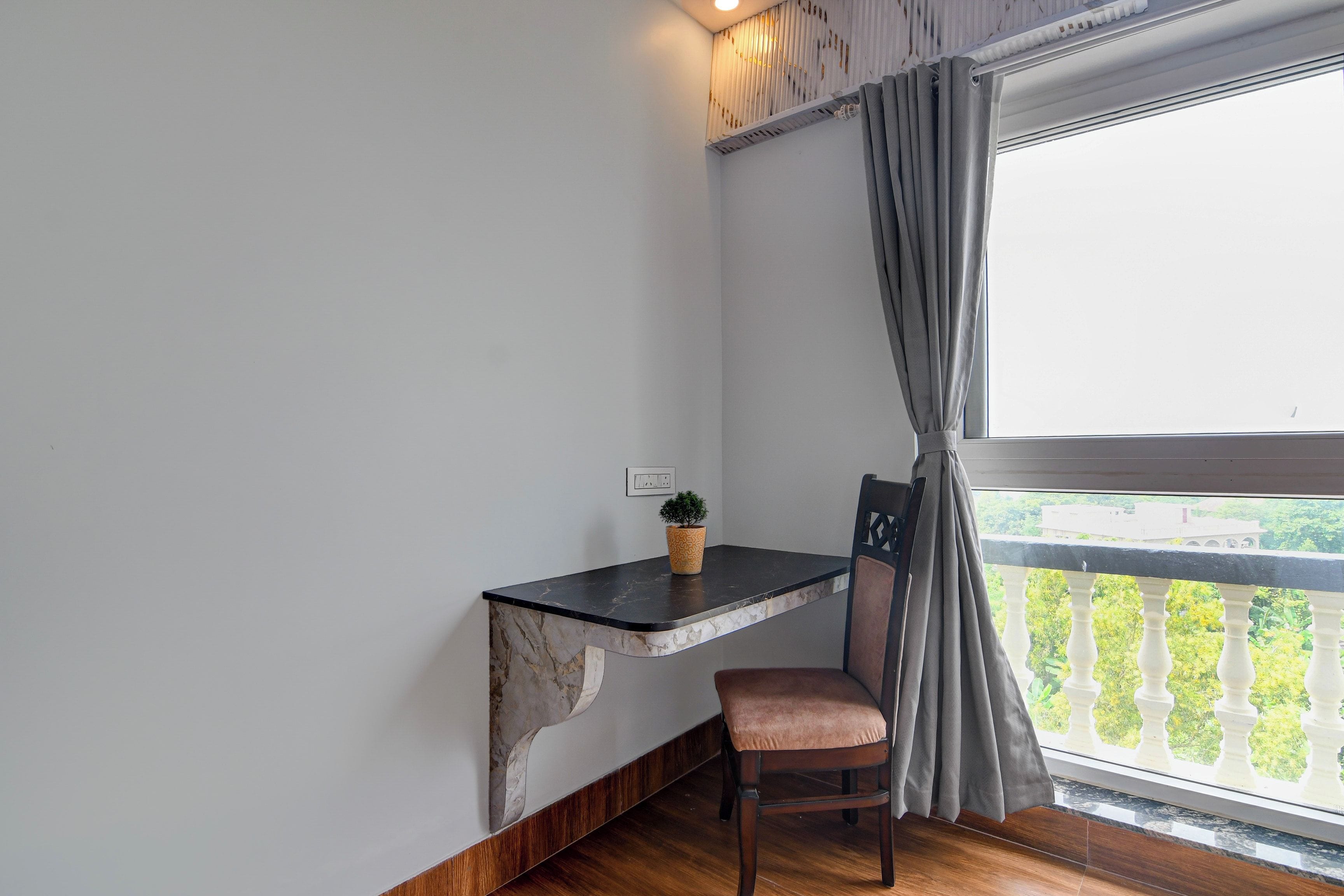 Habitación de esquina con escritorio de mármol, una silla, planta en maceta y vista a la vegetación desde un balcón.