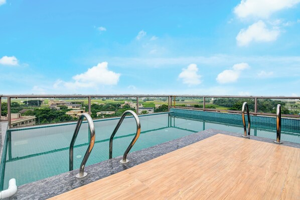 A rooftop infinity pool with a wooden deck and metal railings, overlooking a residential area.