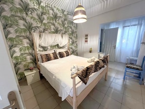 Ein Schlafzimmer mit einem Bett, einem Nachttisch, einem Sessel, einer Pendelleuchte und einem Fenster mit Vorhängen.