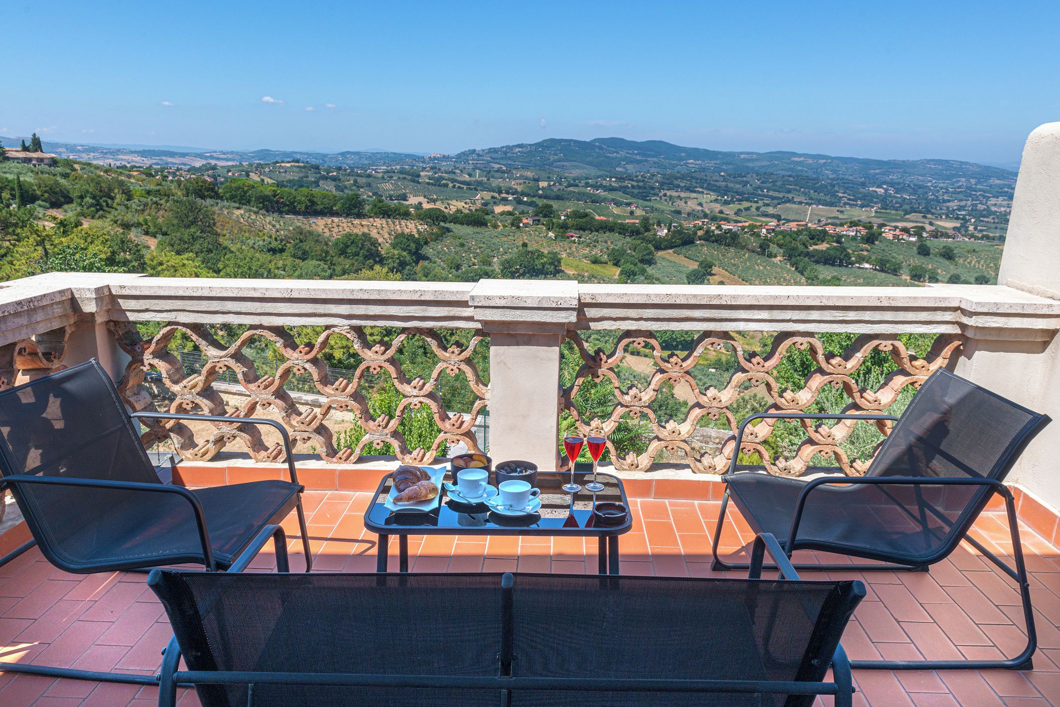 A terrace with two chairs, a table with food and drinks, and a scenic view of hills and vineyards.